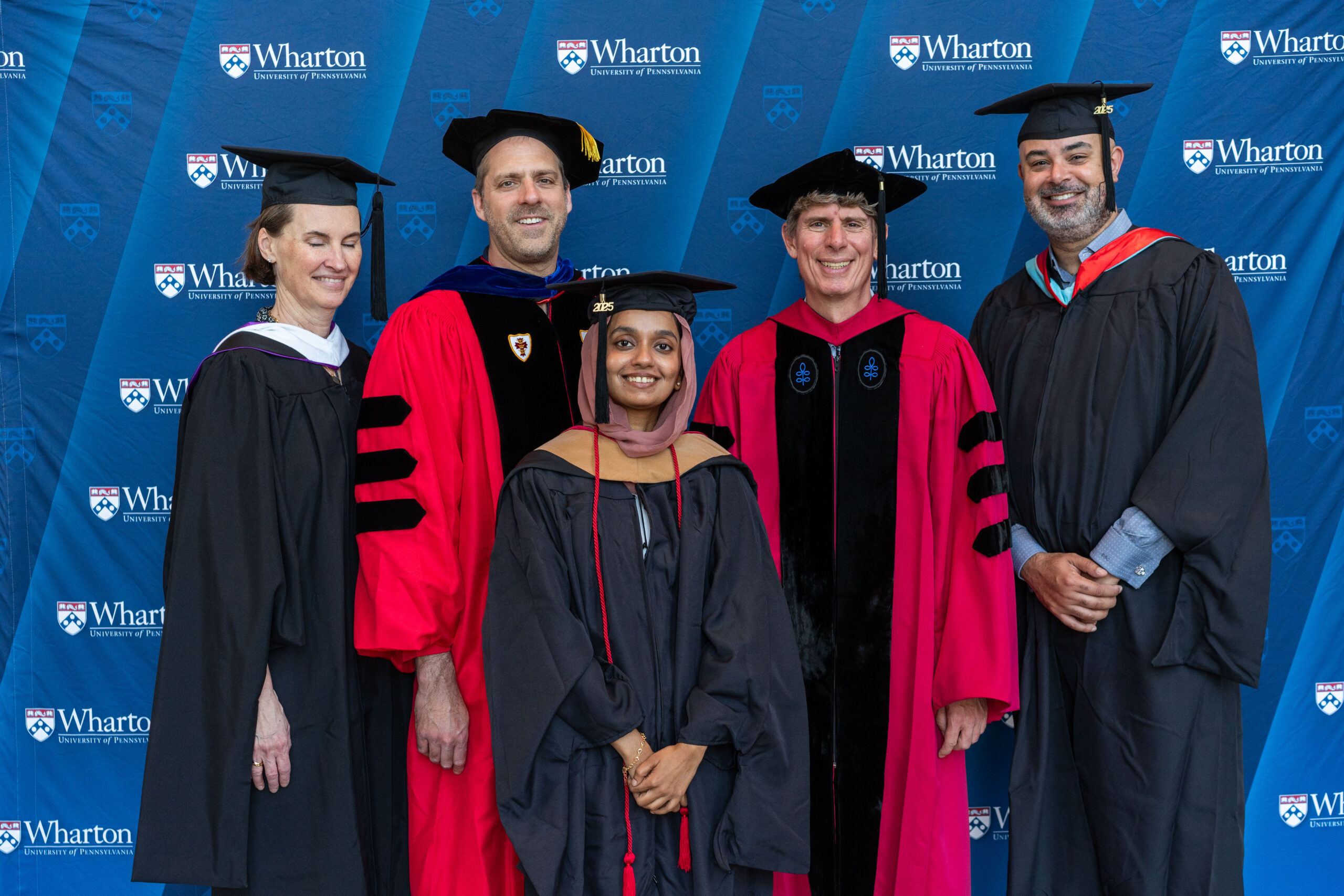 A group of people in academic regalia pose in front of a Wharton School banner. The central figure appears to be a graduate, surrounded by individuals who could be faculty members.