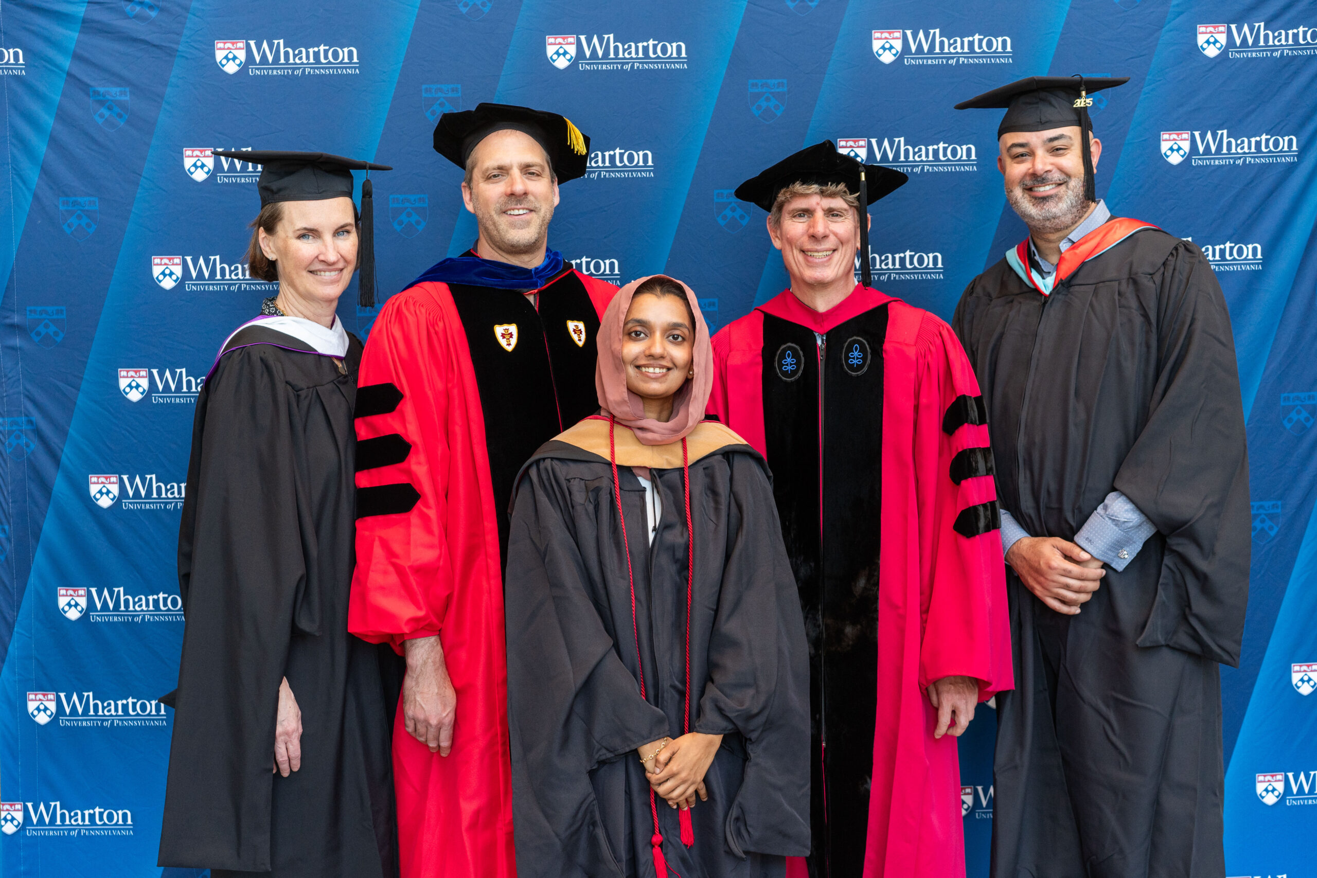 A group of people in academic regalia, including caps and gowns, standing in front of a backdrop with "Wharton" and "University of Pennsylvania" logos.