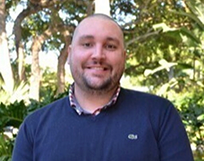 Tripodi Headshot of a person smiling, wearing a dark sweater over a checked shirt, with a background of greenery and trees.